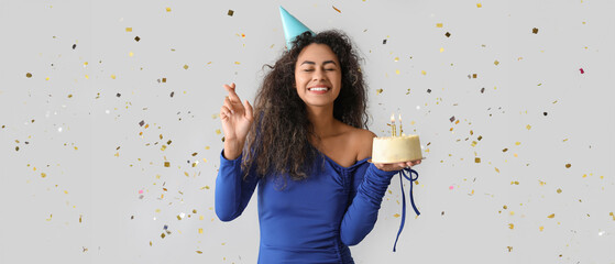 Fototapeta premium Beautiful young African-American woman in party hat with sweet cake and confetti celebrating Birthday on grey background