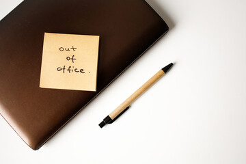 Sticky note with handwritten phrase out of office on a laptop with pen top view