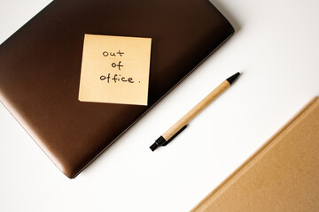 Sticky note with handwritten phrase out of office on a laptop with pen and notebook top view
