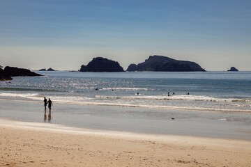 Couple walks along a sunny beach, surfers in the ocean, scenic coastal view. Matauri Bay, Matauri Bay, Northland, NZ