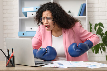 Shocked African-American businesswoman with boxing gloves at table in office. Knockout concept