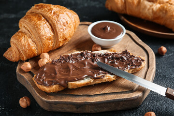 Wooden board of sweet croissants with chocolate spread and hazelnuts on black background