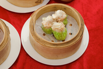 A bamboo steamer contains vibrant green dumplings topped with delicate shrimp, set against a red backdrop.