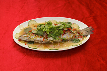 A beautifully presented whole fish dish garnished with herbs and vegetables, served on a decorative plate against a vibrant red background.
