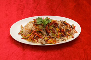 A vibrant dish featuring colorful stir-fried vegetables, rice, and garnishes, served on a white plate against a rich red backdrop.
