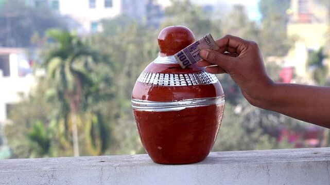 A woman's hand carefully places a 1000 Taka banknote of Bangladesh onto a clay bank, with a serene outdoors background. This image symbolizes the concept of saving money for future needs.
