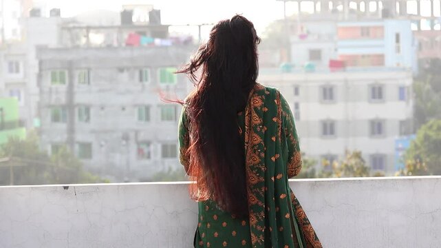 A Bengali woman, wearing a green block-printed salwar kameez, stands alone on a rooftop. Her long, reddish-black hair is gently moved by the breeze as she gazes out, lost in thought. Back view.