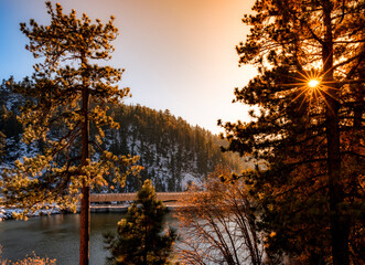 Sun shining through a pine tree with snowy mountainside background with lake and bridge in the foreground 