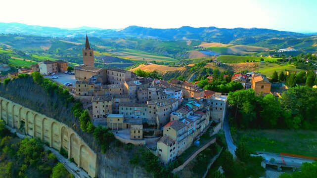 Phenomenal aerial pullback shot from Castignano (AP), camera receding to capture the entire town center perched on a rugged hill, buildings ascending to the spired bell tower, among Marche landscapes