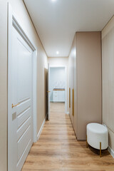 There is a long and spacious hallway featuring elegant wooden floors and a stylish white ottoman placed thoughtfully in the corner, adding a touch of sophistication to the area