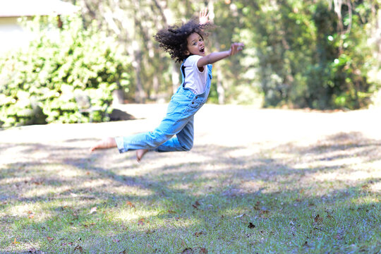 A joyful child in overalls leaps mid-air outdoors on a sunny day