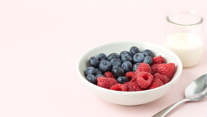 Close view of raspberries and blueberries in a white bowl, metal spoon and a small glass of milk on the side, on light pink background with space for text, banner