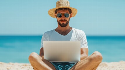 A man with sunglasses and a straw hat works on a laptop while sitting on a beach, enjoying the sunny weather and ocean view.