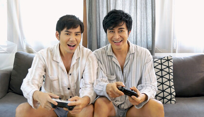 Happy gay couple enjoy playing game together sitting sofa at home living room lifestyle. Friendship gamer men use joystick playing have fun happy time. Two best friends LGBTQ relation partner home