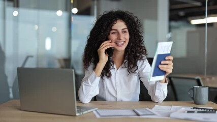Cheerful businesswoman talking on phone and holding passport with tickets in modern office. Curly haired woman in white shirt sitting at table with laptop, excited about her upcoming trip.