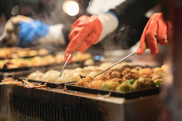 Street vendor making takoyaki with precise tools, steam rising from the hot grill under bright market lighting.