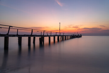 Obraz premium Flat Calm. Long exposure photograph of a wooden pier jutting out to sea at sunrise. The sea is silky smooth and the sky is orange and blue. Ao Cho, Koh Samet, Thailand.