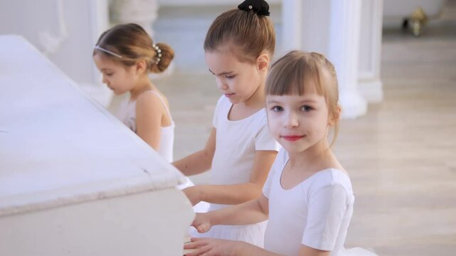 Three young girls are sitting on a piano bench, playing the piano. The girls are wearing white outfits and are smiling. Scene is happy and cheerful