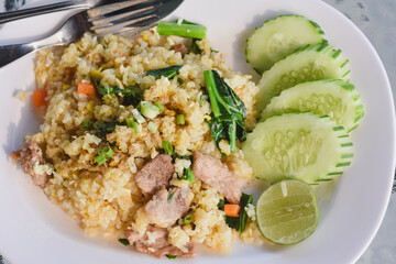 plate of fried rice (Khao Pad) served with slices of cucumber and a wedge of lime on the side.The fried rice contains pieces of meat,likely pork,along with vegetables such as carrots and green onions