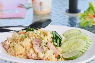 plate of fried rice (Khao Pad) served with slices of cucumber and a wedge of lime on the side.The fried rice contains pieces of meat,likely pork,along with vegetables such as carrots and green onions