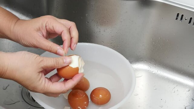 Hands Carefully Peeling Boiled Eggs Over a Bowl of Water in a Kitchen Sink, Showcasing a Simple Cooking Process with Fresh Ingredients and Clean Preparation in a Home Kitchen Setting