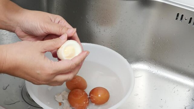 Hands Carefully Peeling Boiled Eggs Over a Bowl of Water in a Kitchen Sink, Showcasing a Simple Cooking Process with Fresh Ingredients and Clean Preparation in a Home Kitchen Setting