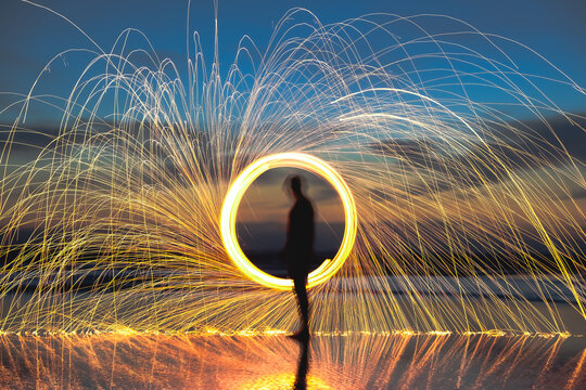 Light trail photo of a ring of fire or spinning sparkler above a water reflection with a colorful backdrop of dusk colors and the ocean.