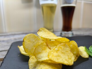 Crispy potato chips are laid out on a board with glasses of beer in the background