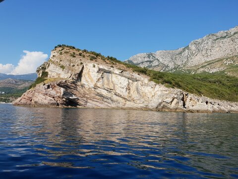 A rocky coastal cliff covered with greenery, surrounded by calm blue water and mountain views under a clear sky