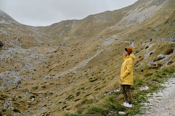 Breathtaking mountain landscape with young traveler woman wearing yellow raincoat. Side view. Travel around the world concept. Montenegro country, Durmitor National Park. Saddle Pass in autumn season.