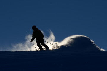 A backlit skier is sliding down a mountain covered in snow and ice, throwing up cascades of snow with a blue sky in the background