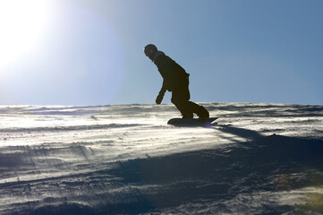 A backlit snowboard rider is sliding down a mountain covered in snow and ice with a blue sky in the background