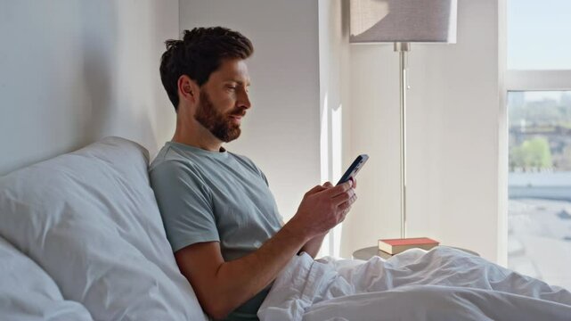 Awake guy scrolling smartphone in bed early morning. Relaxed brunette messaging