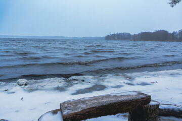 Fototapeta premium Snow-covered lakeshore with wooden bench and table on a winter day