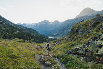 Fototapeta premium A female hiker with a backpack walks along a rugged trail in the Pyrenees, Spain, surrounded by mountains, greenery, and a distant lake at sunset