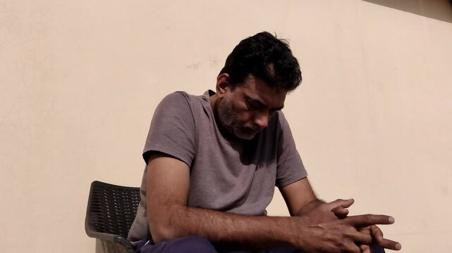 A worried man with a small white shave on his face and black hair sits anxiously on a chair under the bright sun. He gazes downward nervously moving his fingers reflecting a moment of deep concern.