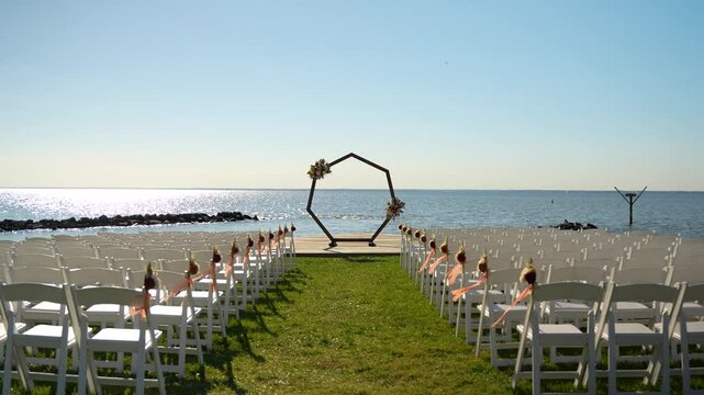 A shot symmetrically glides down a ceremony aisle lined with white chairs adorned with orange ribbons on lush grass. A heptagon centerpiece, draped in flowers, stands framed by blue skies and water.