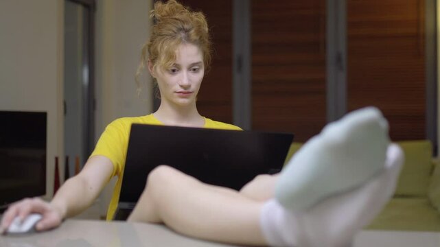 Girl working on a laptop in her lap. Casual clothing with feet wearing different color socks up on the table in a living room at home. Portrait of a student taking online course. Home education.