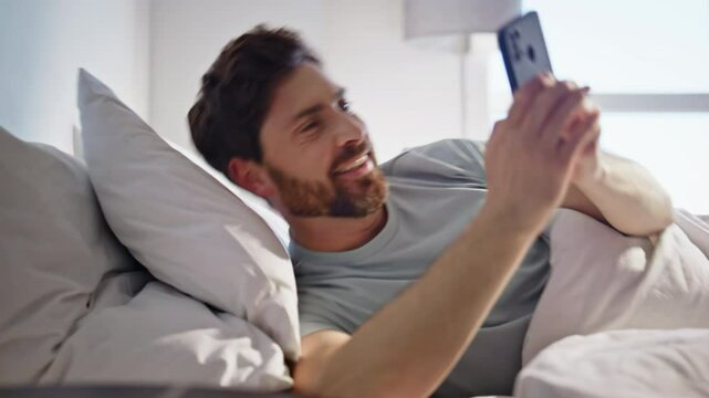 Happy guy looking smartphone screen in bed closeup. Man lying with mobile phone
