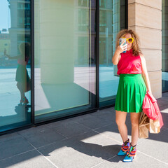 a beautiful young woman in bright clothes with a phone and bags takes selfies or takes pictures of the city on a walk. the concept of fashion, youth, mobile photography, tourism
