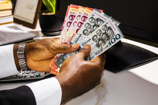 Young black african  man wearing suit sitting at office desk  holding and counting  Ghanaian cedi notes. front desk
