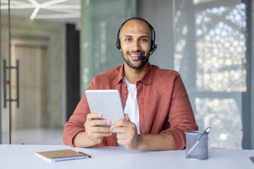 A man with a headset and a tablet computer in his hands looks into the camera, a support service worker at work, advising clients. Web camera view.