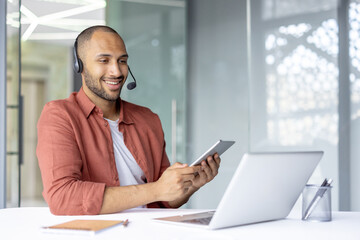 Man with headset working inside office, using tablet computer and laptop. Support service worker at...