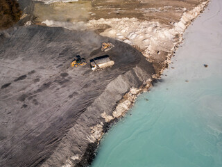 A cement factory tailings site with heavy machinery operating near a turquoise lake. Trucks and...