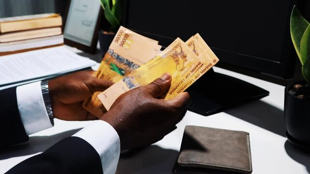 Black African male business man hands wearing suit sitting and counting Ugandan shilling notes at office on desk