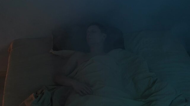 A man lies in bed, coughing and suffocating due to smoke from a house fire, surrounded by a dangerous atmosphere.