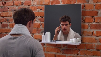 Young man in the morning stands in front of the mirror in bathroom with towel over neck, doing...