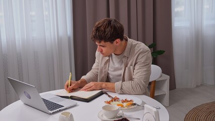 Young man in the morning in the kitchen dinning room enjoying healthy breakfast, sitting at table working typing on laptop and writing notes in notebook.