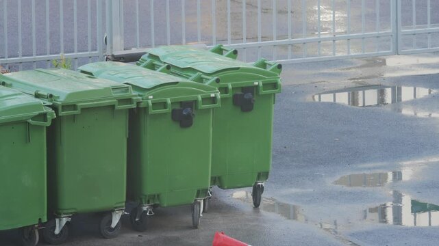Row of large movable rigid plastic green trash bins used to contain unsorted municipal solid waste. Green bins on cloudy rainy day.