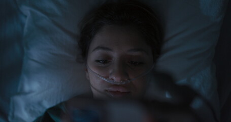 Female patient wearing oxygen tube in hospital bed holds phone in hands chats with friends.